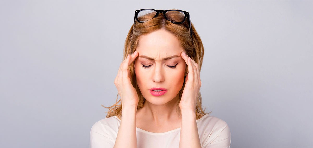 Woman with glasses perched on her head presses her temples and closes her eyes, showing headache discomfort, in a neutral setting.